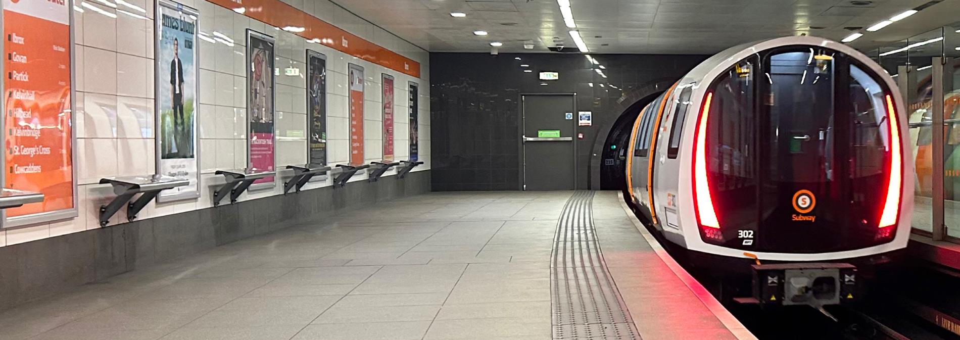 Ibrox station platform with a Subway train departing the station to the right and seats and advertising posters to the left